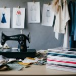 A cluttered room with fabric scraps, sewing machine, and clothing sketches pinned to the wall. A stack of fashion magazines sits on a table